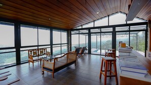 A spacious room with large windows offering a view of the landscape. There are wooden furniture pieces, including a sofa and chairs, and a table with books. - Spice Tree Munnar (Udumbanchola)