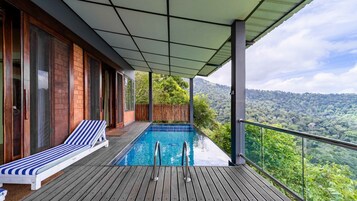 Una terraza de madera con piscina, una silla de descanso y vistas a una cadena montañosa cubierta de bosques.
