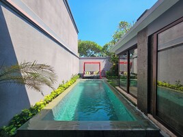 Area piscina esterna moderna con vasca azzurra, tenda rossa e zona relax con cuscini.