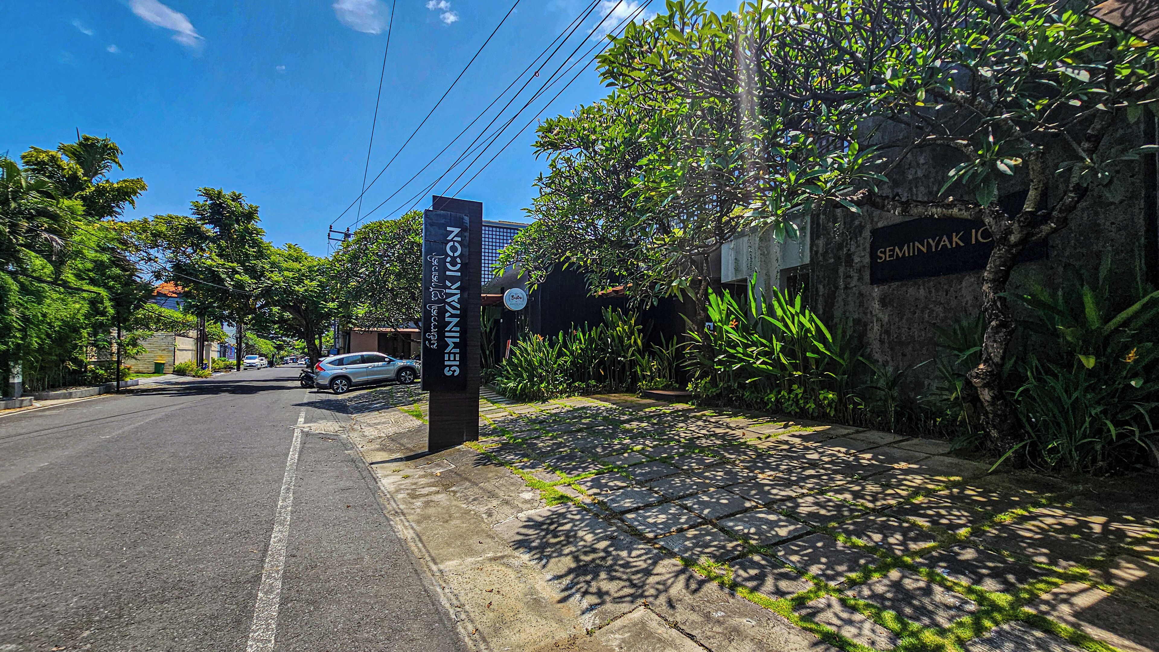 Une vue de rue avec une enseigne indiquant « SEMINYAK ICON » et une autre indiquant « Bistro », le tout entouré d’une végétation luxuriante et sous un ciel bleu dégagé.