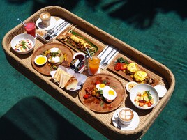 A breakfast tray with various dishes including eggs, toast, and fruit, set on a wicker table by a pool.