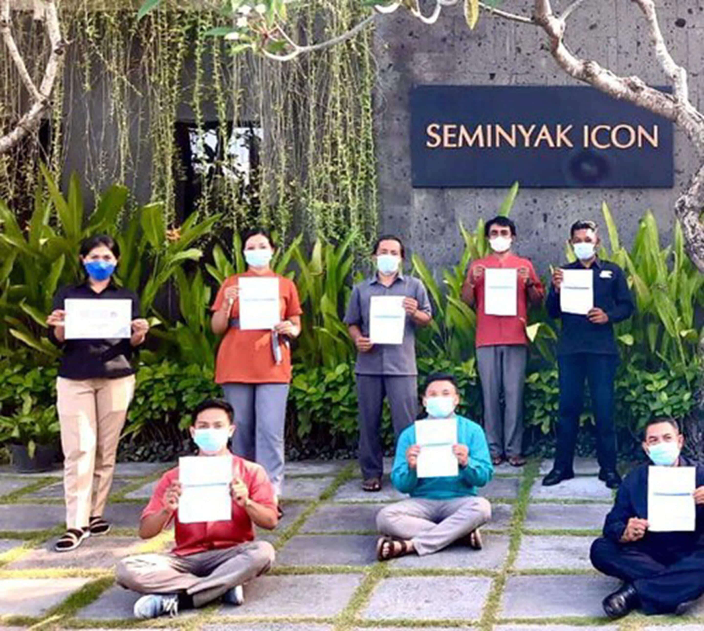 Un grupo de personas sosteniendo certificados frente a un letrero que dice 'SEMINYAK ICON'.
