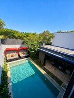 Un área de piscina exterior moderna con una piscina azul cristalina, dos zonas de estar con cubiertas rojas y un muro de piedra.