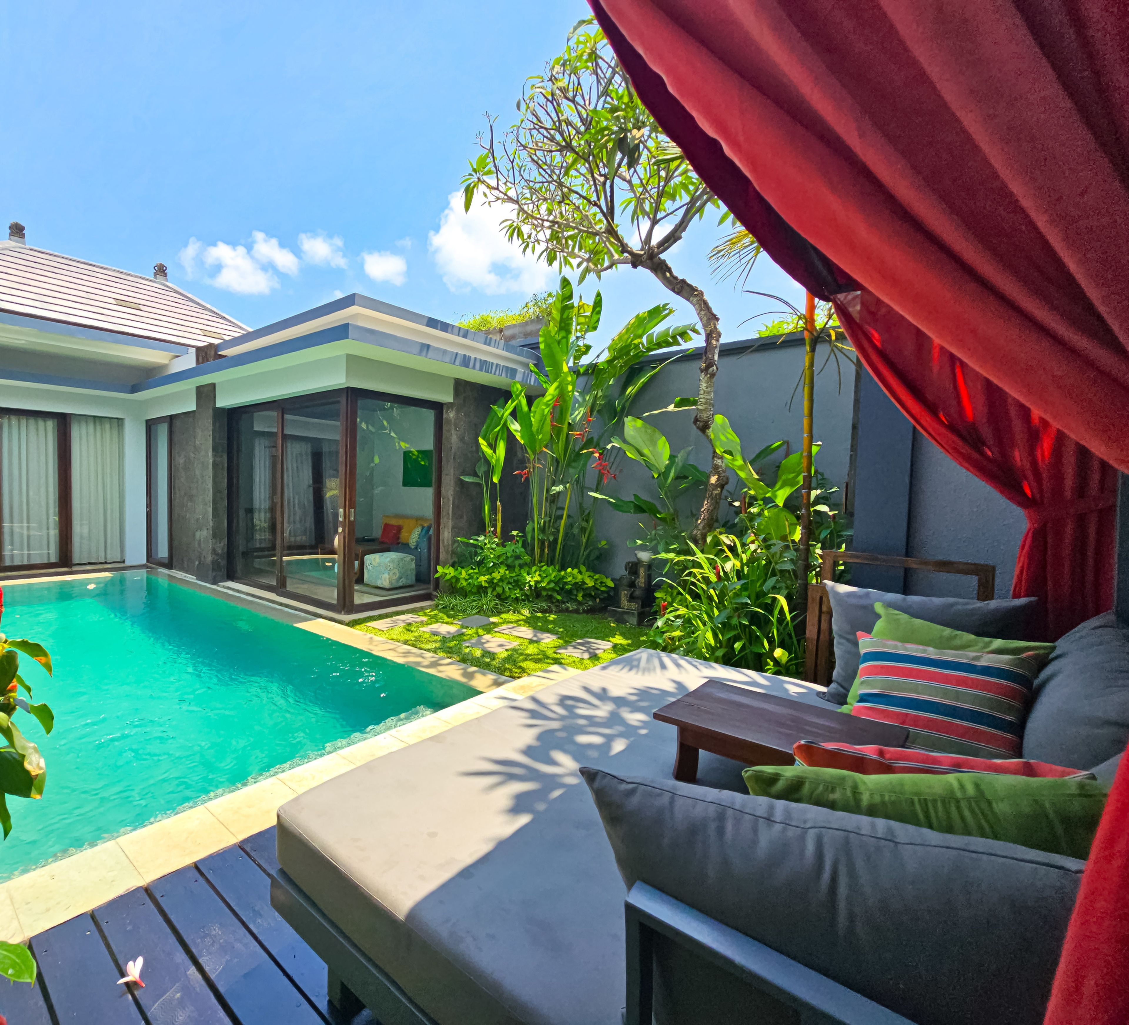A modern outdoor pool area with a glass-enclosed seating area, lush greenery, and a red canopy.