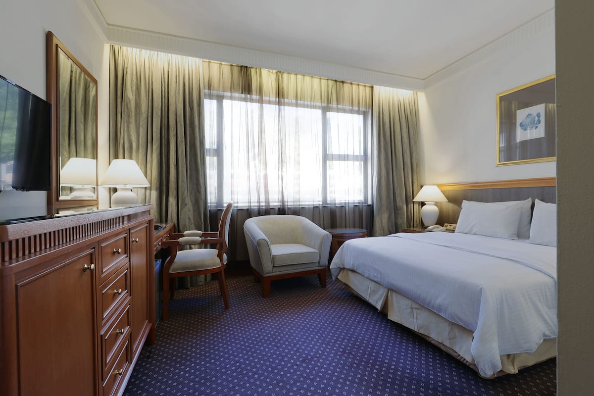 Une chambre d’hôtel avec un lit, un bureau avec une lampe, une chaise, une télévision et une grande fenêtre avec des rideaux.