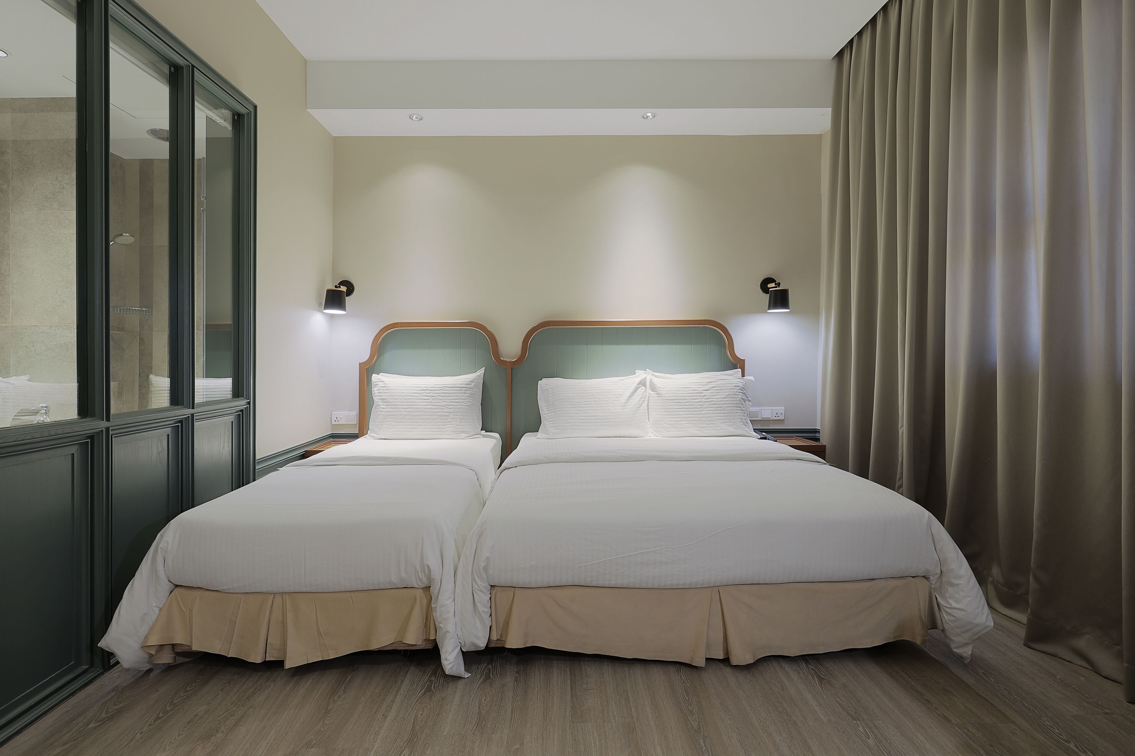 A double bed with a wooden headboard and a beige skirt, two bedside tables, a wall-mounted light, and a window with a view of a building.