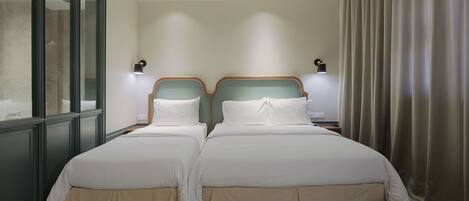 A double bed with a wooden headboard and a beige skirt, two bedside tables, a wall-mounted light, and a window with a view of a building.