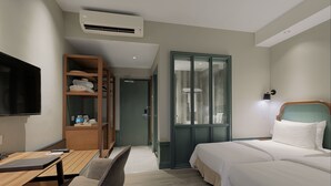 Habitación de hotel con cama, escritorio con silla, televisor, armario y baño con puerta de vidrio.
