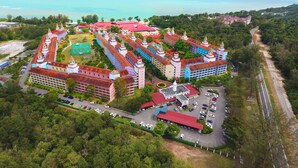 Une vue aérienne d’un grand complexe hôtelier coloré, composé de plusieurs bâtiments, avec un parking et une vue sur l’océan.