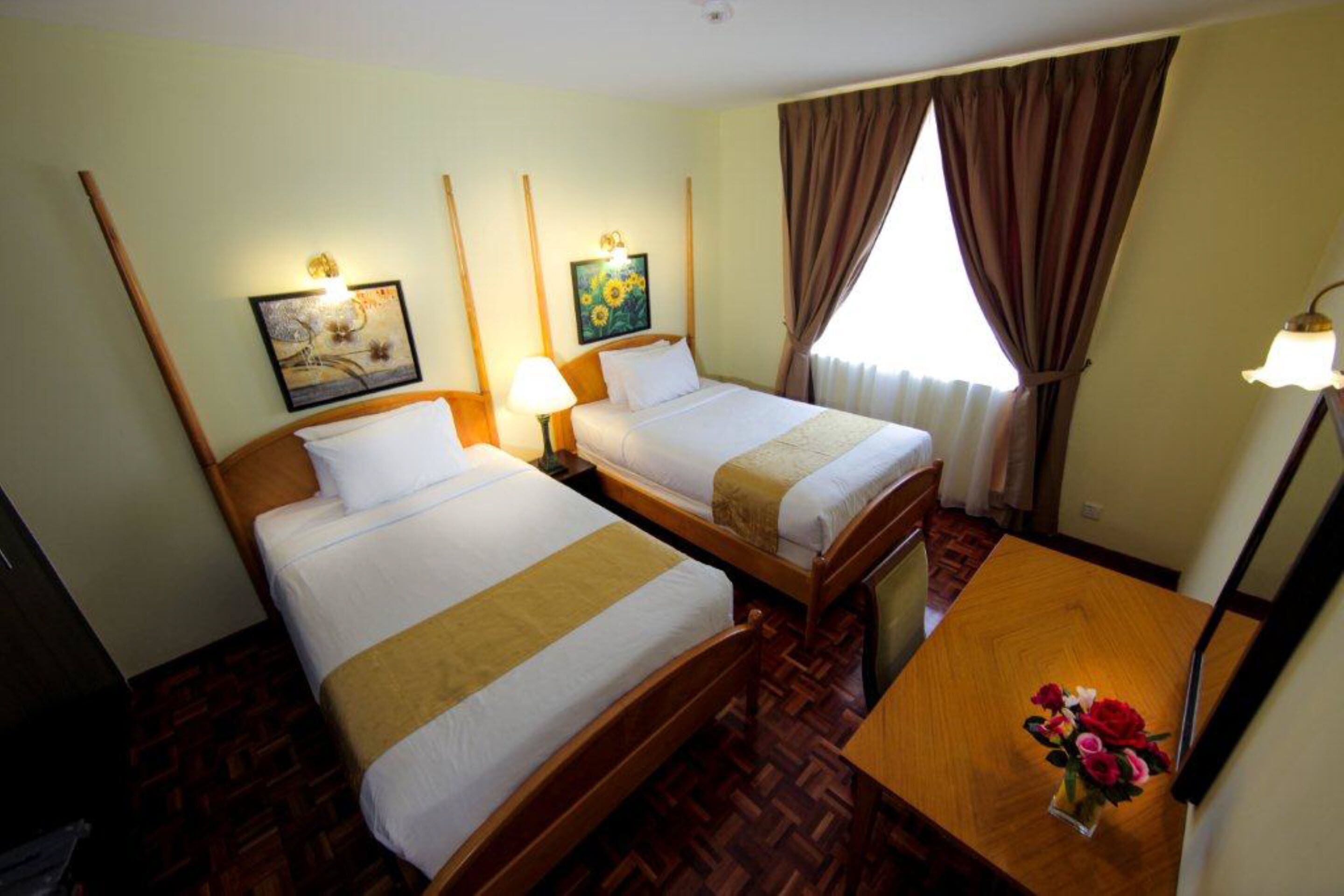 A hotel room with two single beds, a wooden floor, a wooden table with a vase of flowers, and a window with curtains.