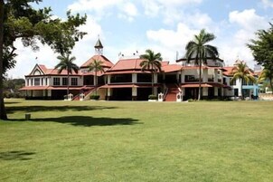 A large, multi-story building with a red roof and white walls, surrounded by a well-maintained lawn and palm trees.
