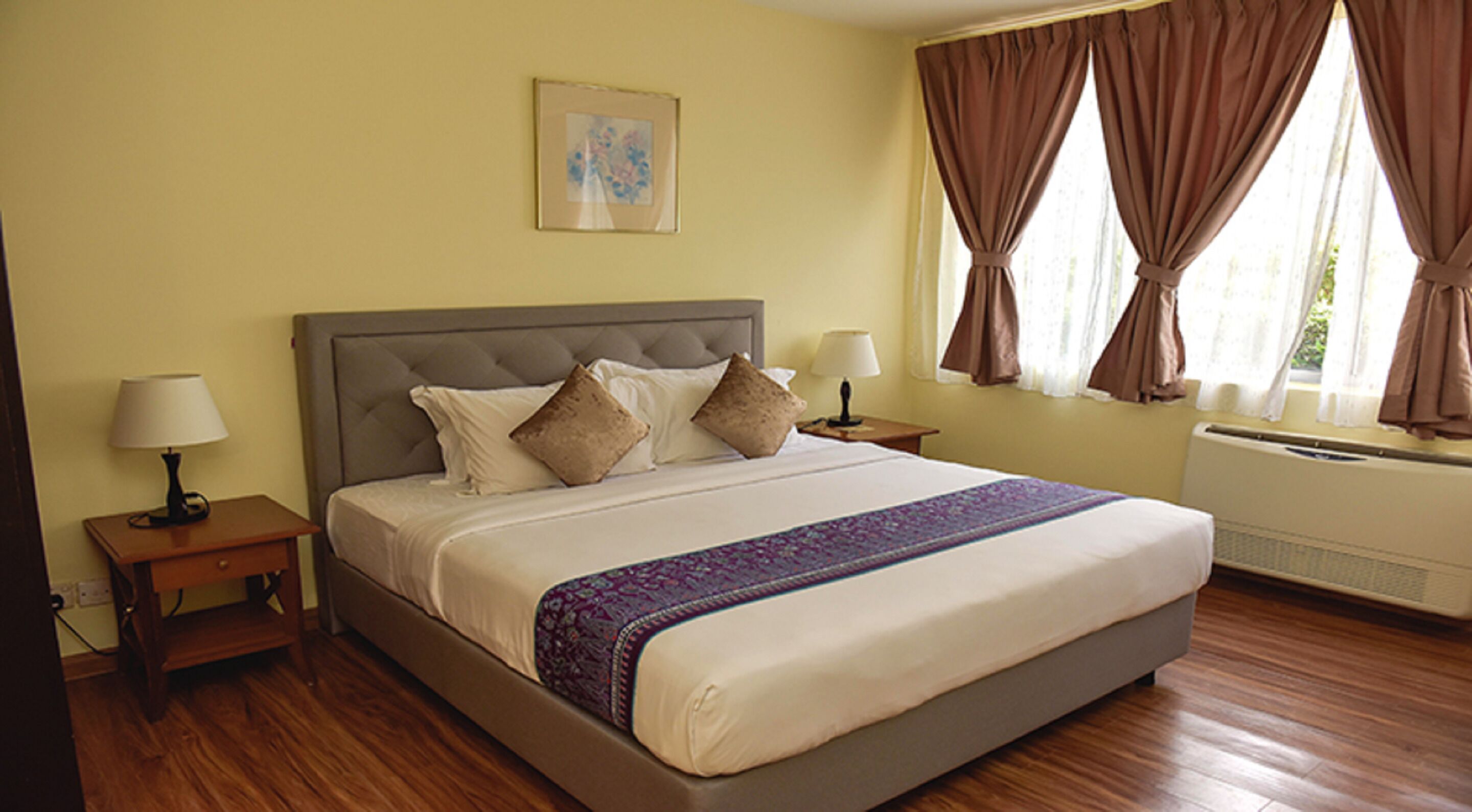 A neatly made bed with a patterned bedspread, two bedside tables with lamps, a wall-mounted picture, and a window with curtains.