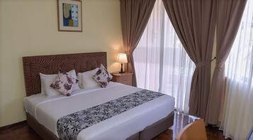 A neatly made bed with a patterned bedspread and pillows, a headboard with a geometric design, a bedside lamp, and sheer curtains on a window.