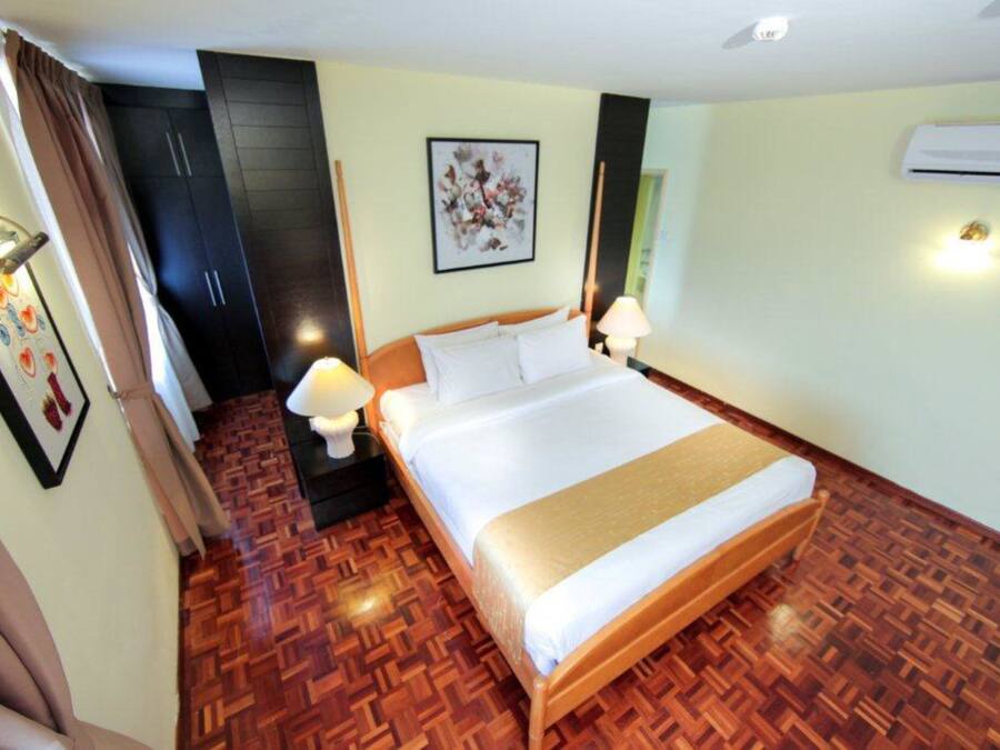 A hotel room with a large bed, wooden flooring, a nightstand with a lamp, a wall-mounted air conditioner, and a framed picture on the wall.