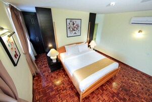 Quarto de hotel com cama grande, piso de madeira, mesa de cabeceira com abajur, ar condicionado fixado na parede e quadro emoldurado.