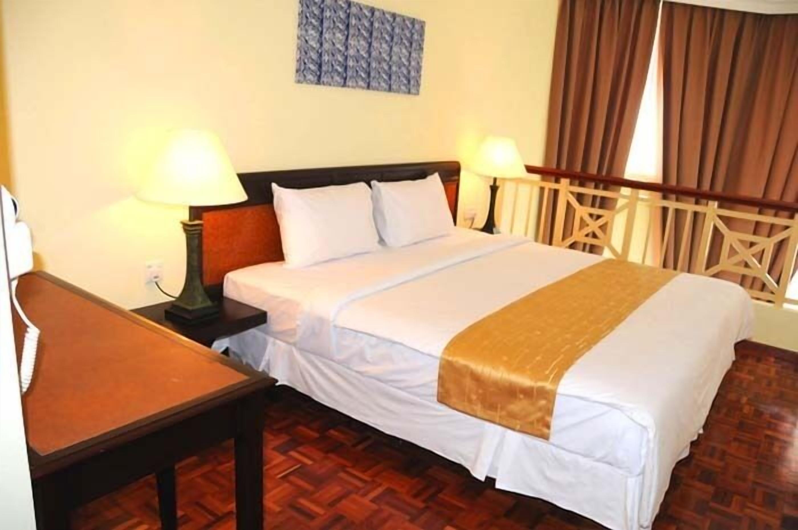 A hotel room with a bed, two bedside lamps, a wooden headboard, a wooden table, and a patterned wall hanging.