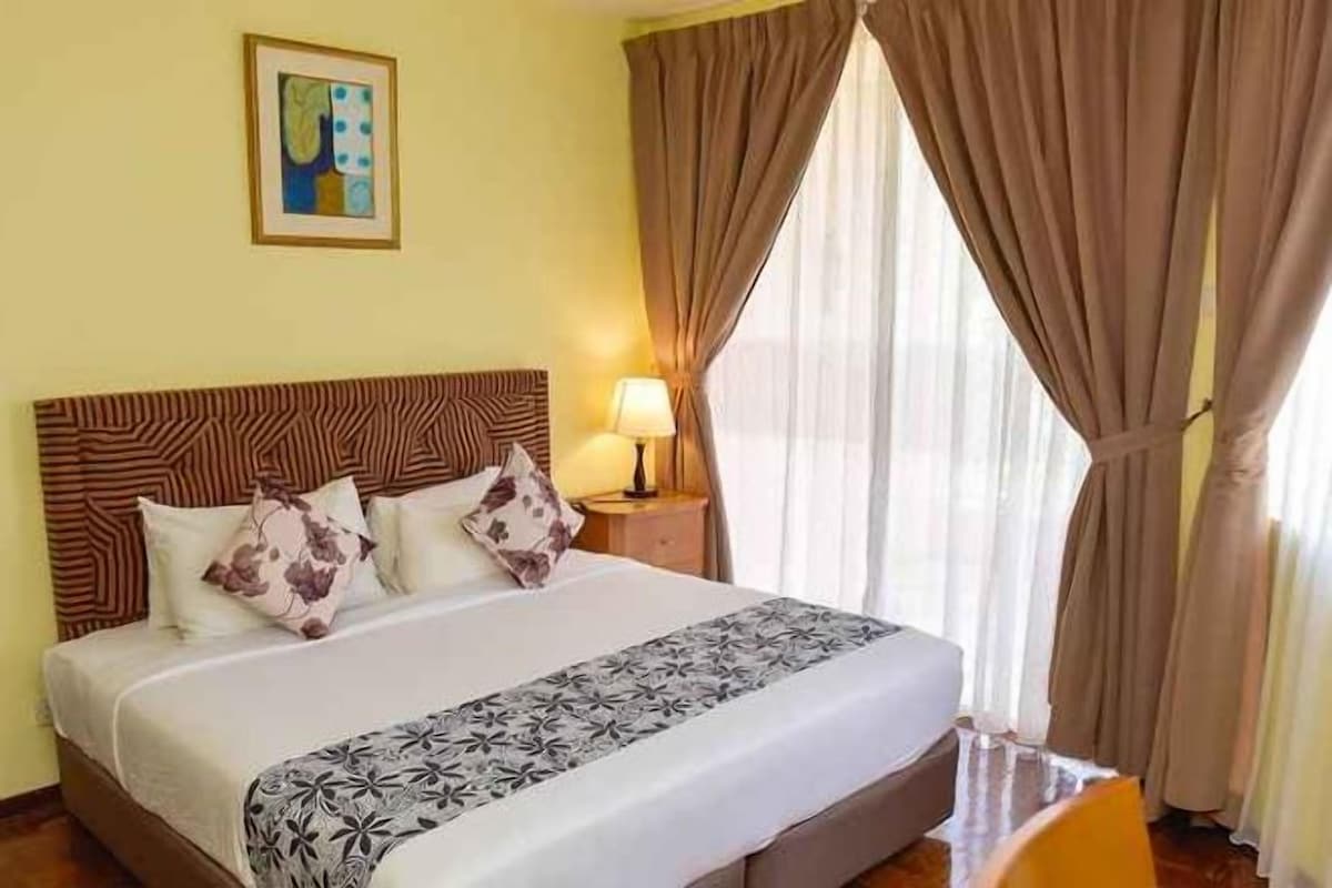 Quarto de hotel com cama, mesa de cabeceira, luminária e janela com cortinas.