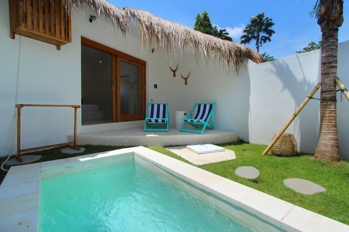 A swimming pool with two striped lounge chairs, a thatched-roof building, and a palm tree.