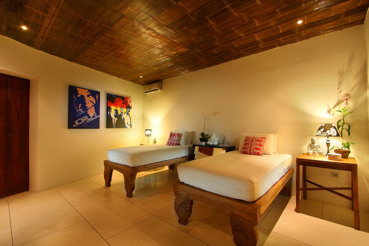 A bedroom with two beds, a wooden ceiling, and a small table with a lamp and plant.