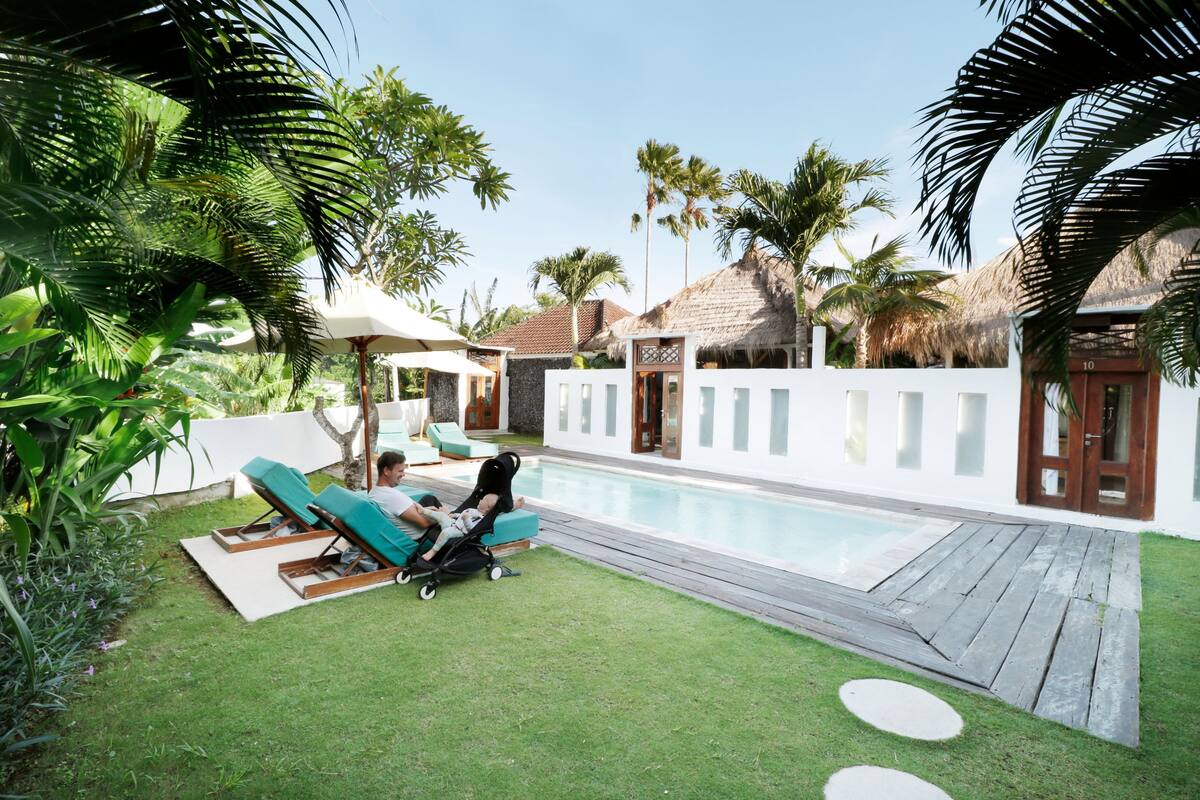 A poolside area with lounge chairs, a baby stroller, and a thatched-roof building.