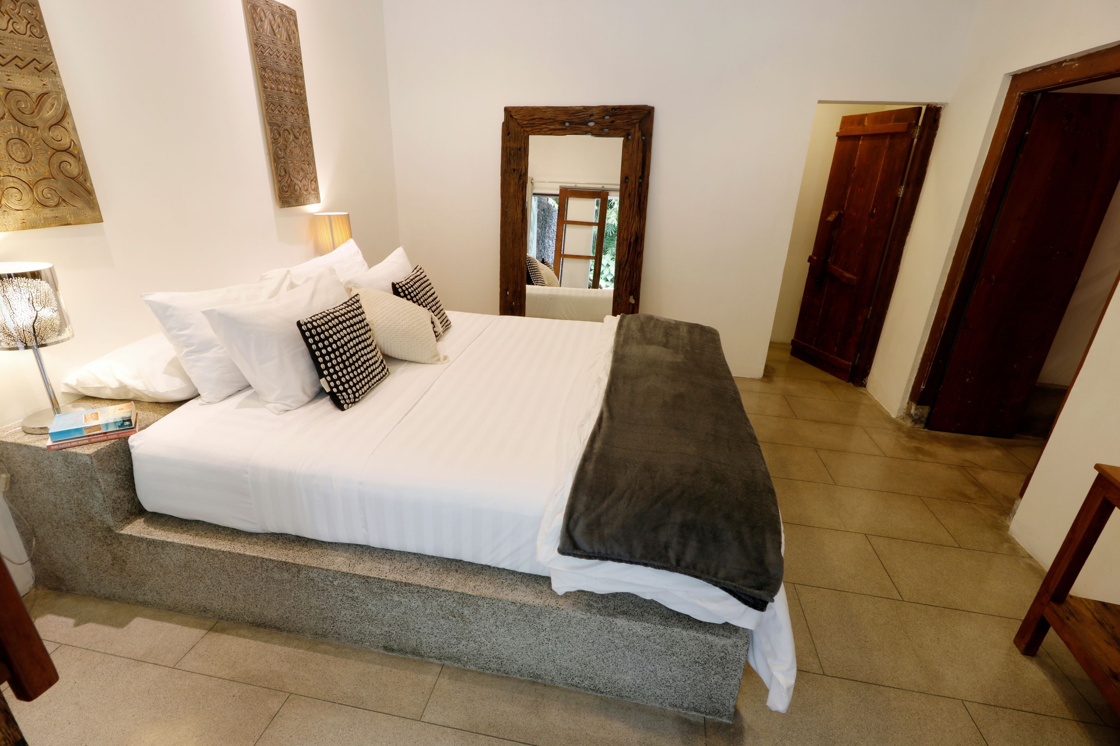 A neatly made bed with white linens and multiple pillows, a bedside lamp, and a framed mirror on the wall.