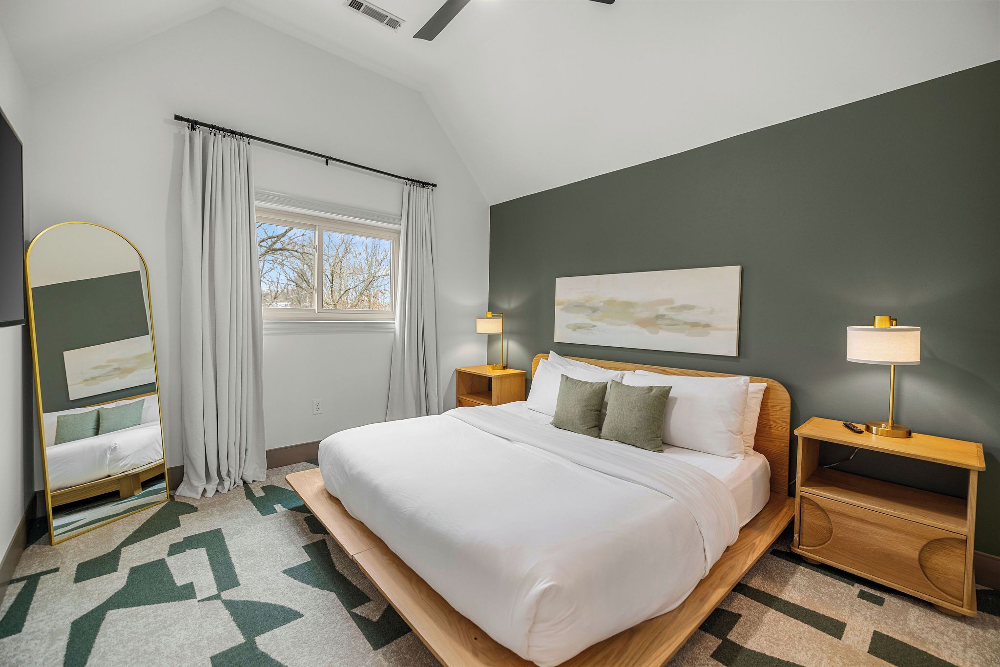 A modern hotel room with a large bed, two bedside tables with lamps, a mirror, a window with curtains, and a geometric patterned rug.