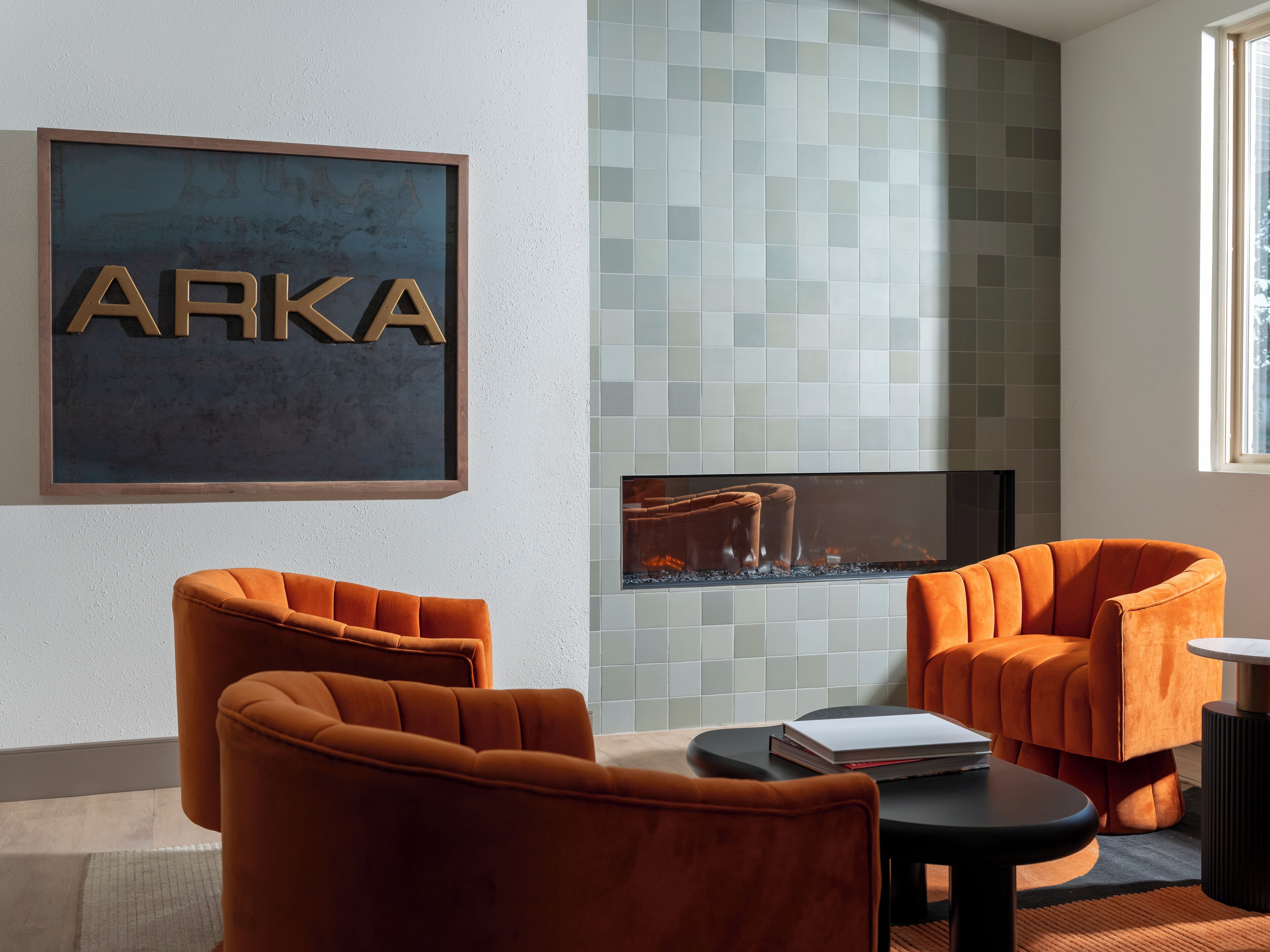 Un intérieur moderne avec une enseigne encadrée portant l’inscription « ARKA », une cheminée agrémentée de carreaux décoratifs et deux fauteuils orange.