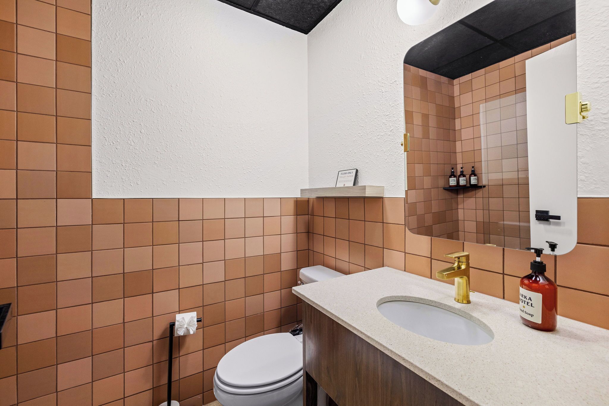 A bathroom with a toilet, sink, and mirror. There is a shelf with toiletries and a sign above the sink.