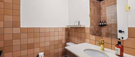 Baño con inodoro, lavamanos y espejo. Se observa una repisa con artículos de aseo y un cartel sobre el lavamanos.