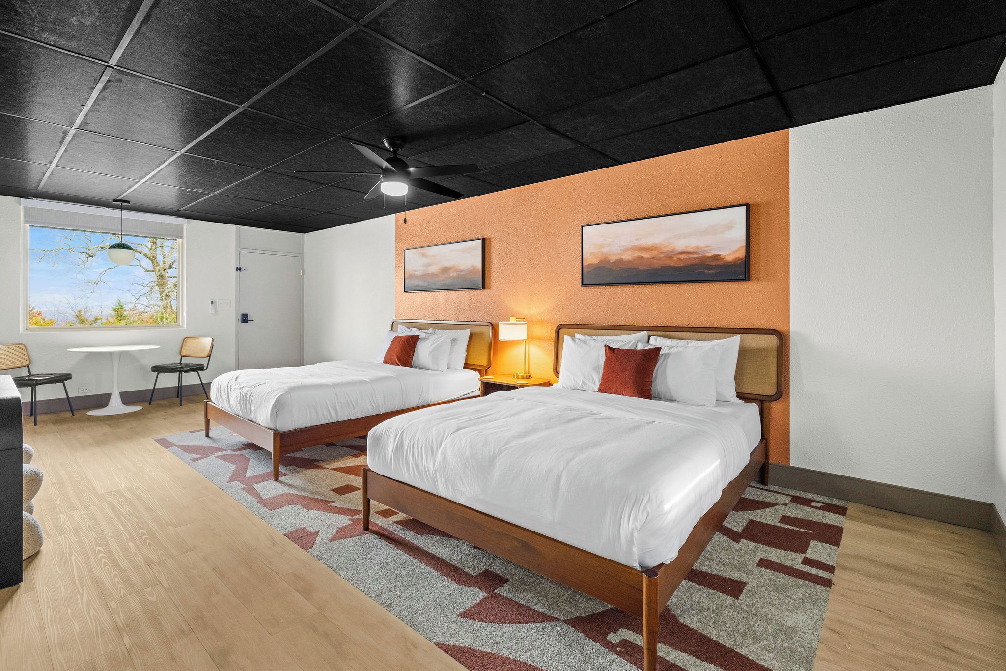 Habitación de hotel con dos camas, un escritorio, sillas, un ventanal con vistas y obras de arte en las paredes.