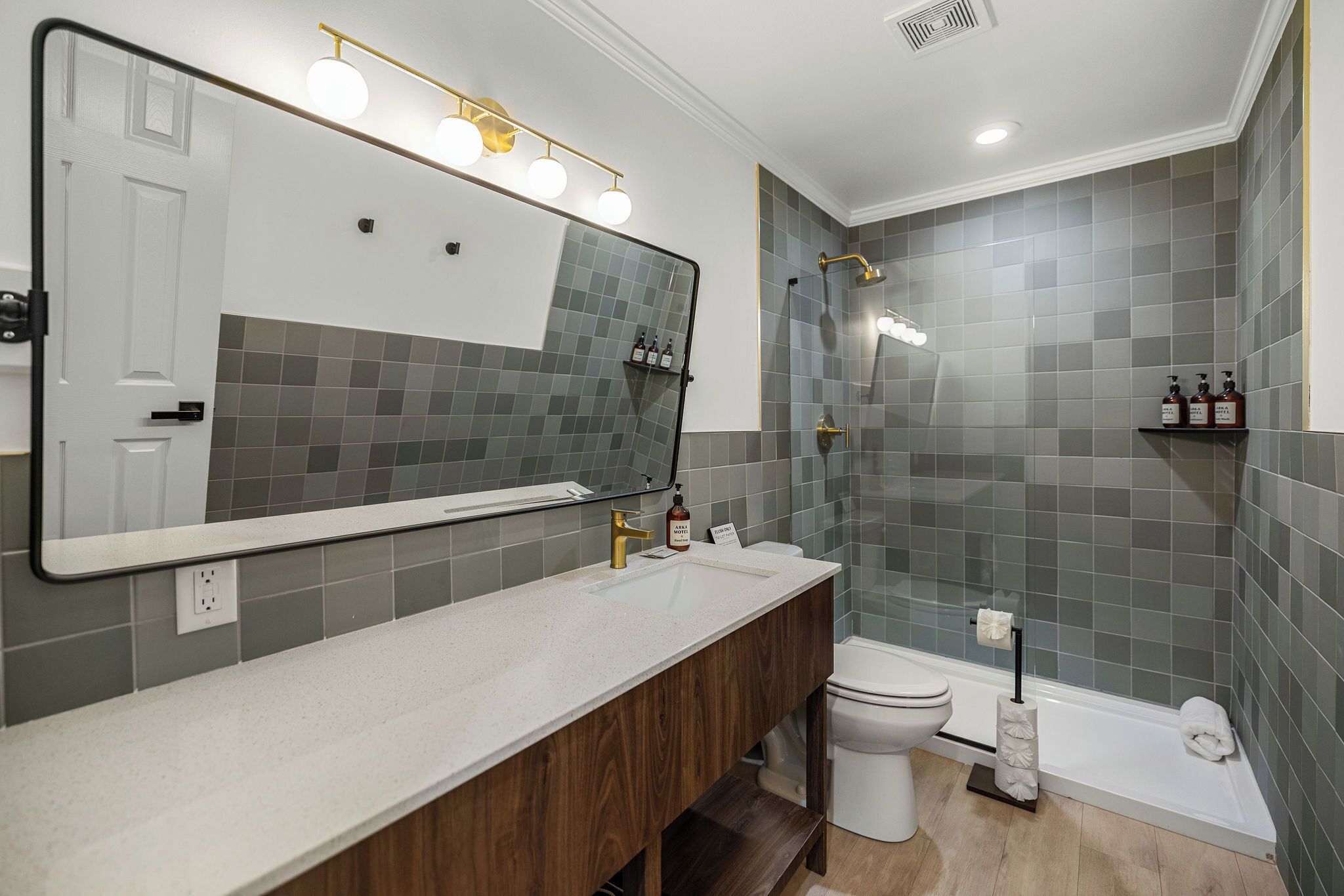 A modern bathroom with a large mirror, a wooden vanity, a shower, and grey tiled walls.