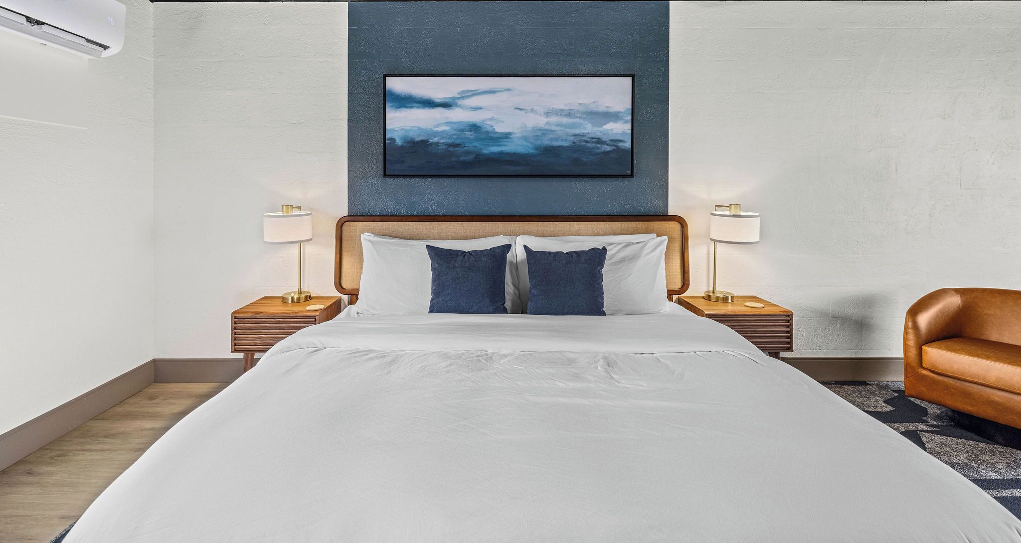 A hotel room with a large bed, two bedside tables with lamps, a brown leather chair, and a framed picture on the wall.
