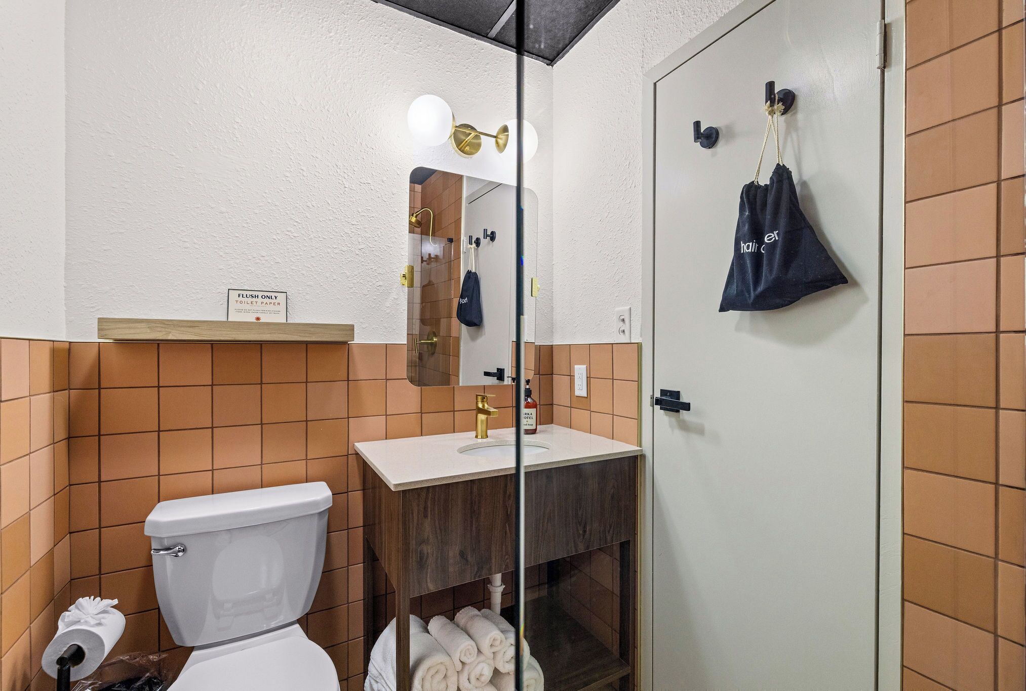 A bathroom with a toilet, sink, and mirror. There is a sign above the toilet and a towel rack with a bag on the wall.