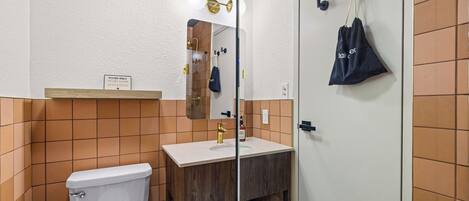 A bathroom with a toilet, sink, and mirror. There is a sign above the toilet and a towel rack with a bag on the wall.