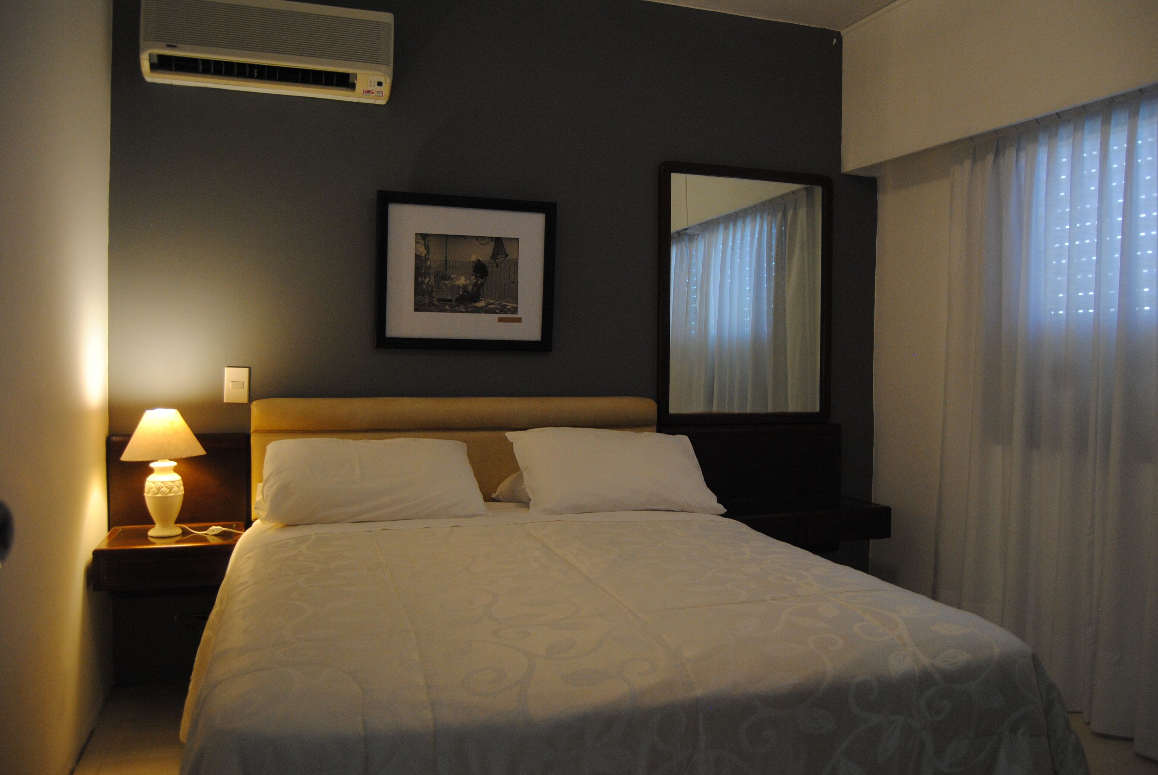 A hotel room with a bed, a nightstand with a lamp, a framed picture on the wall, and a window with curtains.