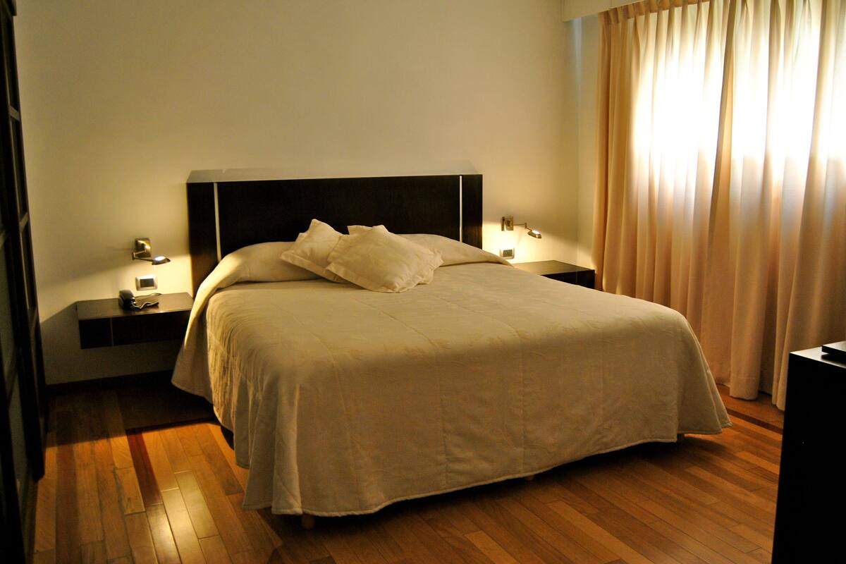 A neatly made bed with a headboard, two bedside tables with lamps, and a wooden floor.