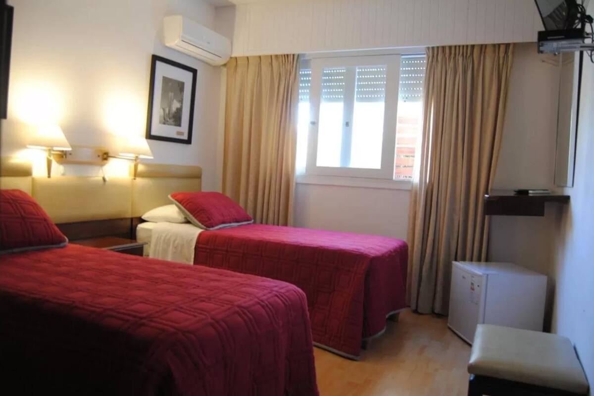 A room with two beds, a small table, a desk, a chair, a window with curtains, and a wall-mounted air conditioner.