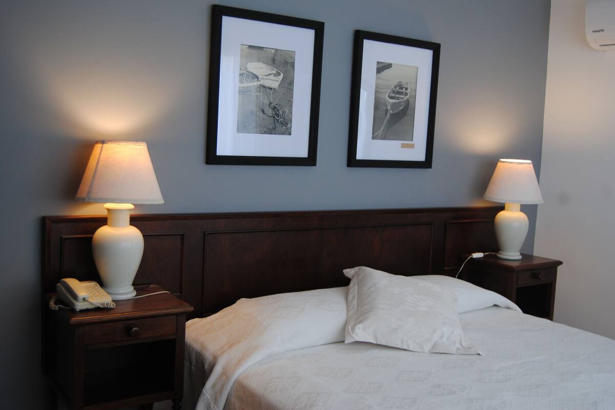 A neatly made bed with white linens, two bedside lamps, a nightstand with a phone, and two framed pictures on the wall.