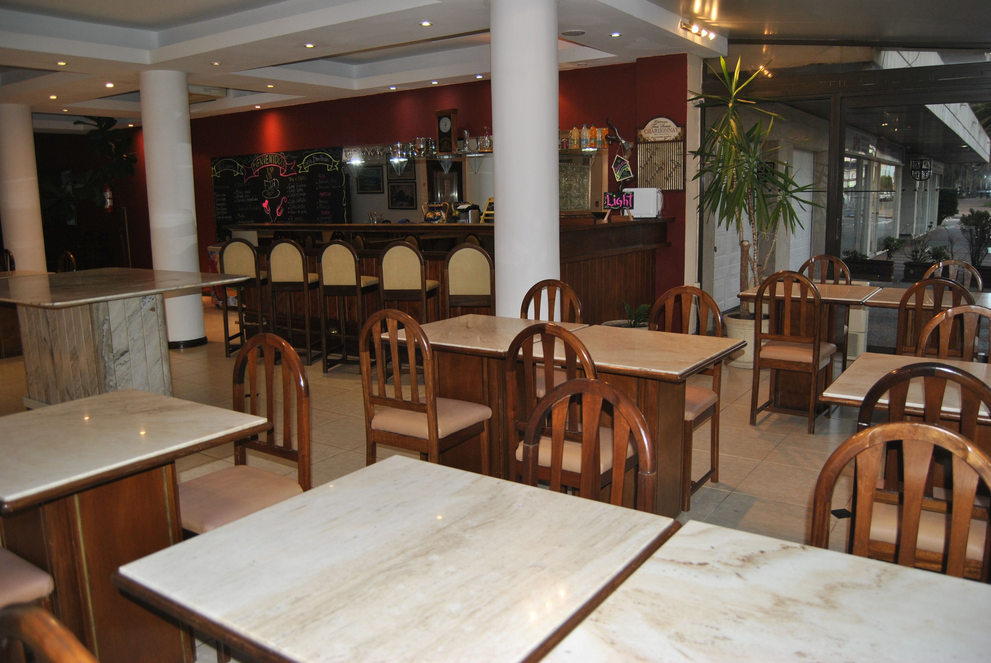 Un restaurante con mesas de mármol, sillas de madera, una zona de bar con estantes llenos de botellas y una planta grande en maceta.