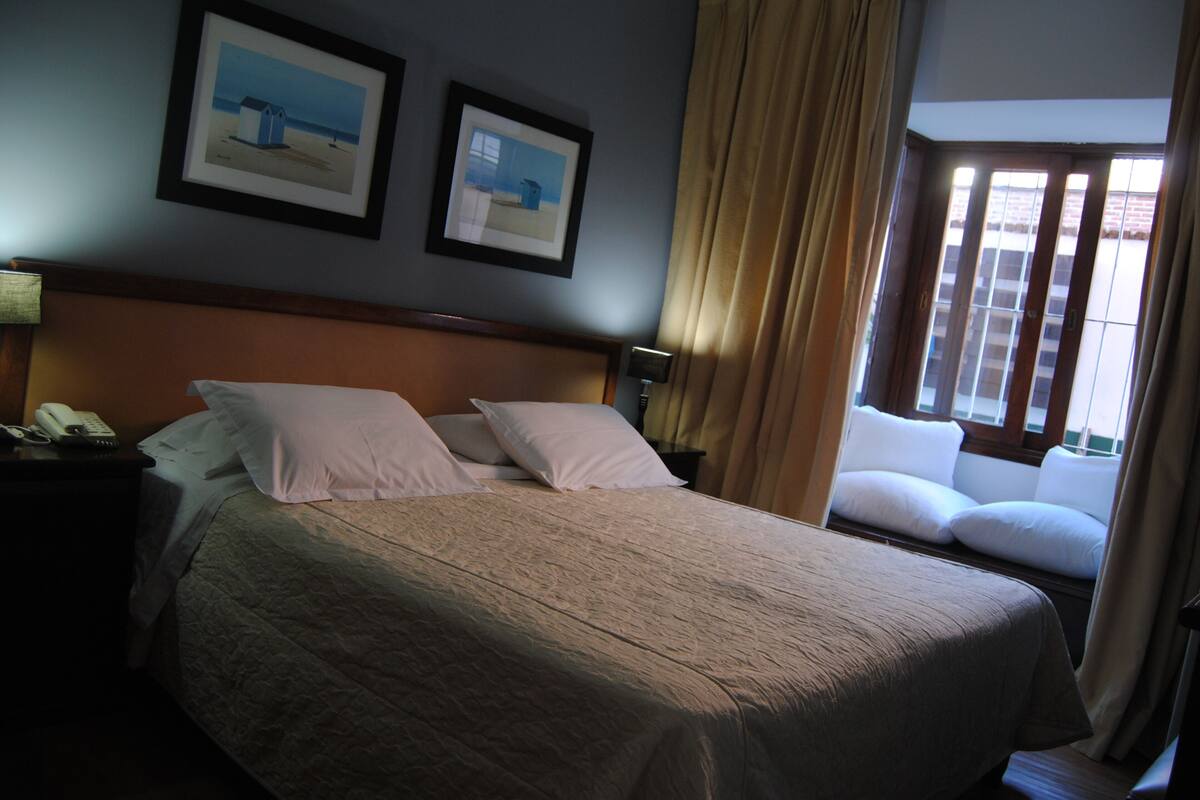 A hotel room with a large bed, two framed pictures on the wall, a bedside lamp, and a window with curtains.