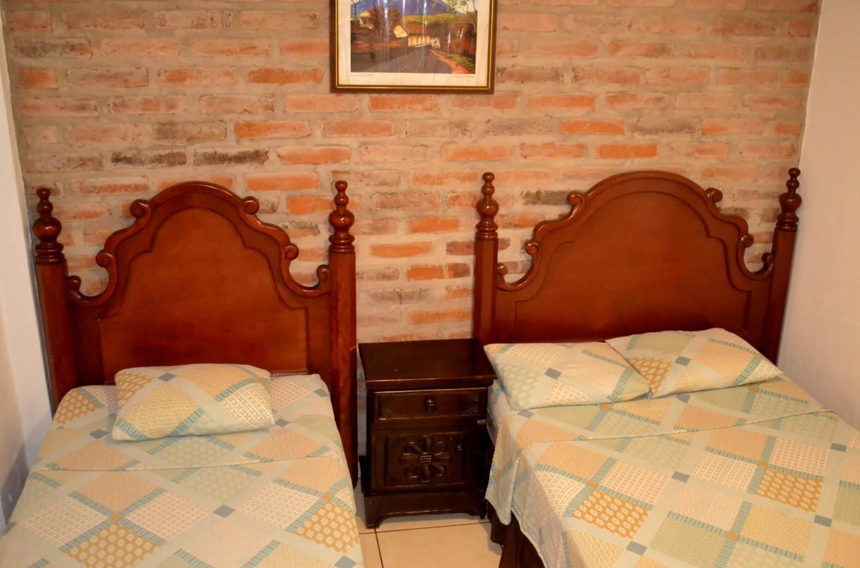 A room with two wooden beds, a small nightstand, and a brick wall with a framed picture.