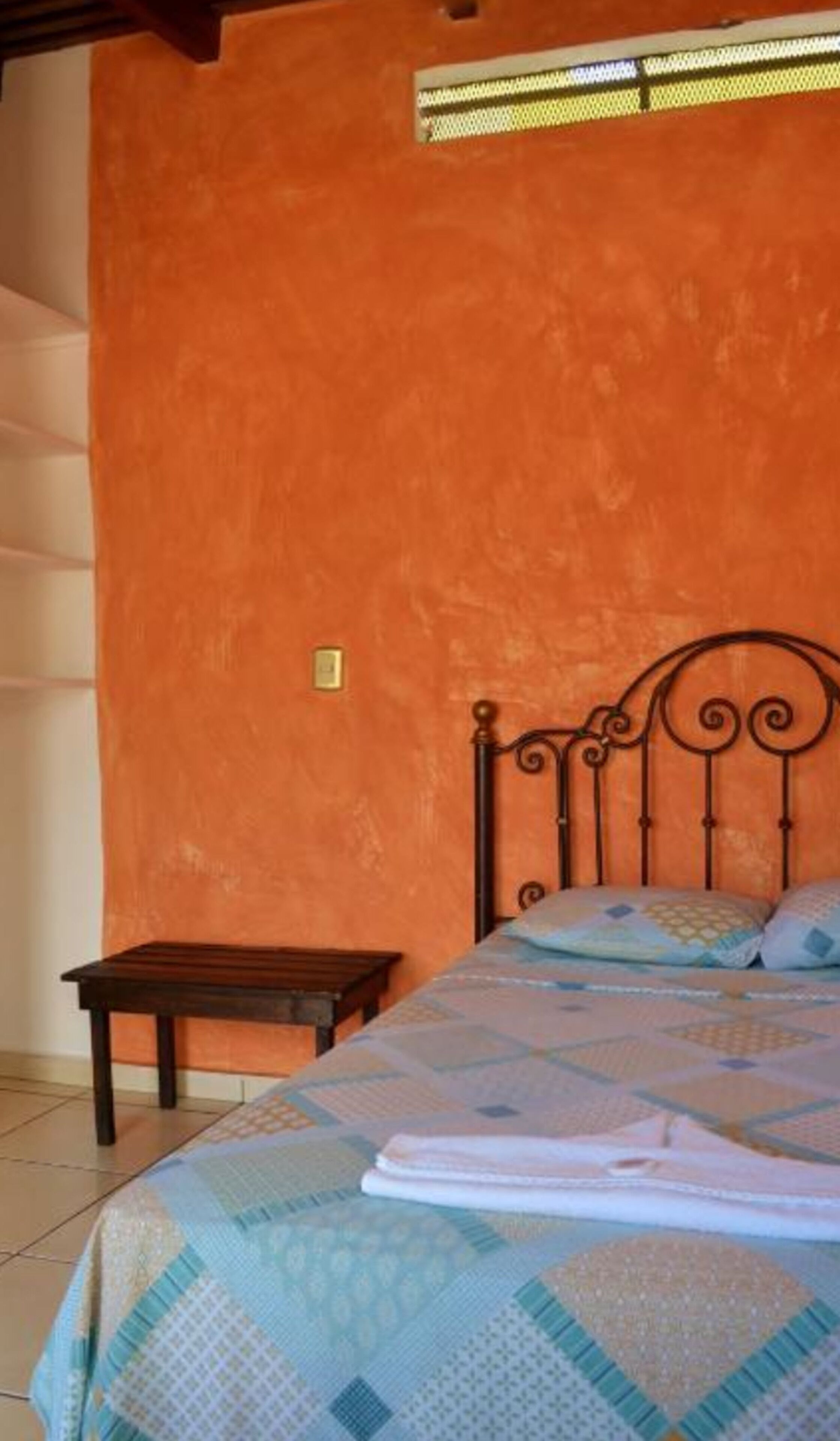 A bedroom with an orange wall, a metal bed frame, a wooden nightstand, and a patterned bedspread.