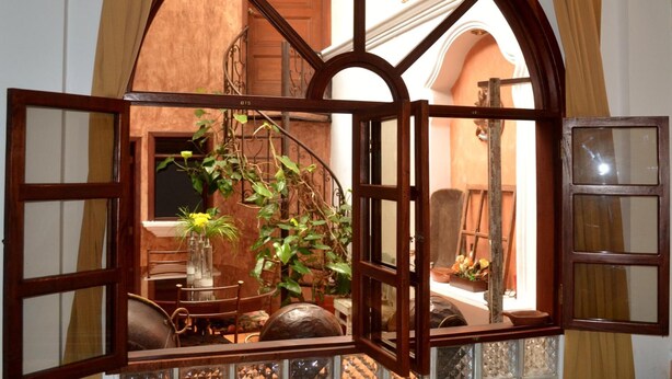 A room with a large arched window, a dining area with a table and chairs, and a staircase visible through the window. - Hotel Villa Florencia Centro (San Salvador)