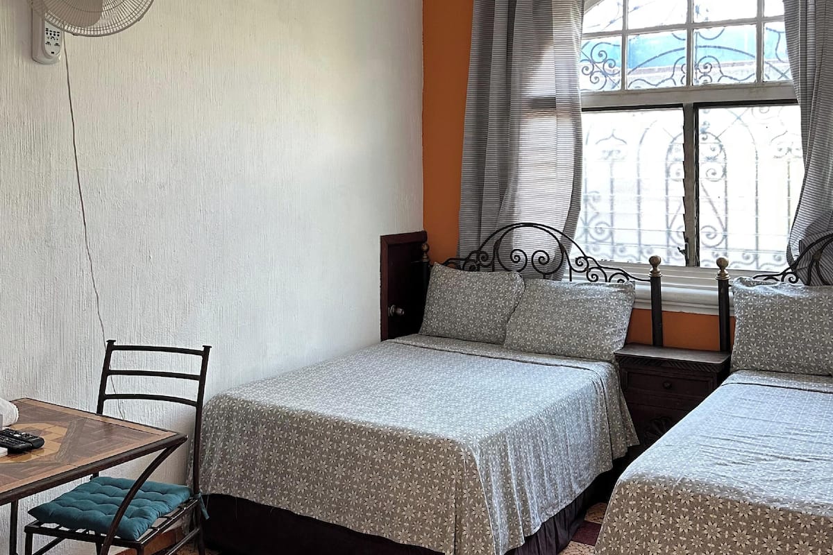 A room with two beds, a table, a chair, a fan, and a window with curtains.