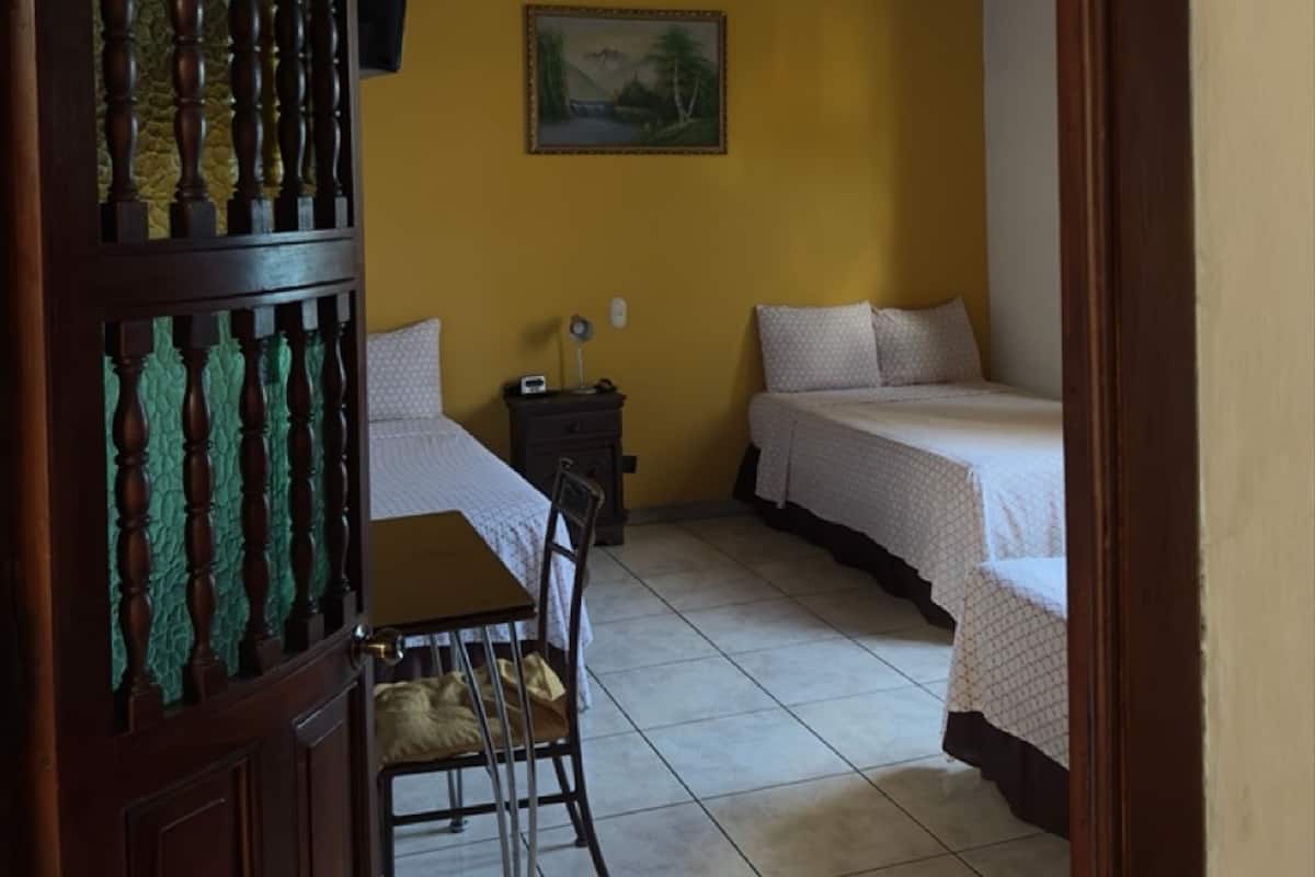 A room with two beds, a wooden chair, a table, a nightstand, and a painting on the wall.
