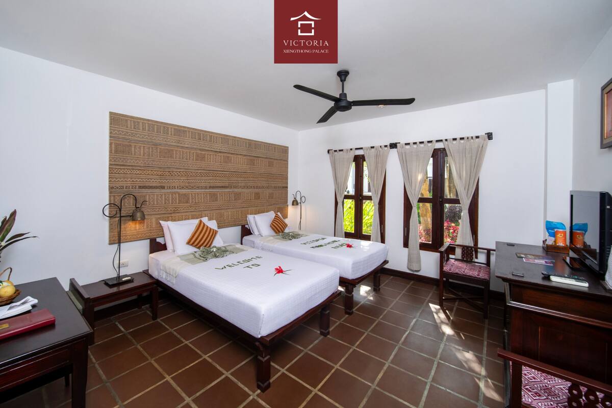 A hotel room with two beds, a desk, a chair, a TV, and a ceiling fan.