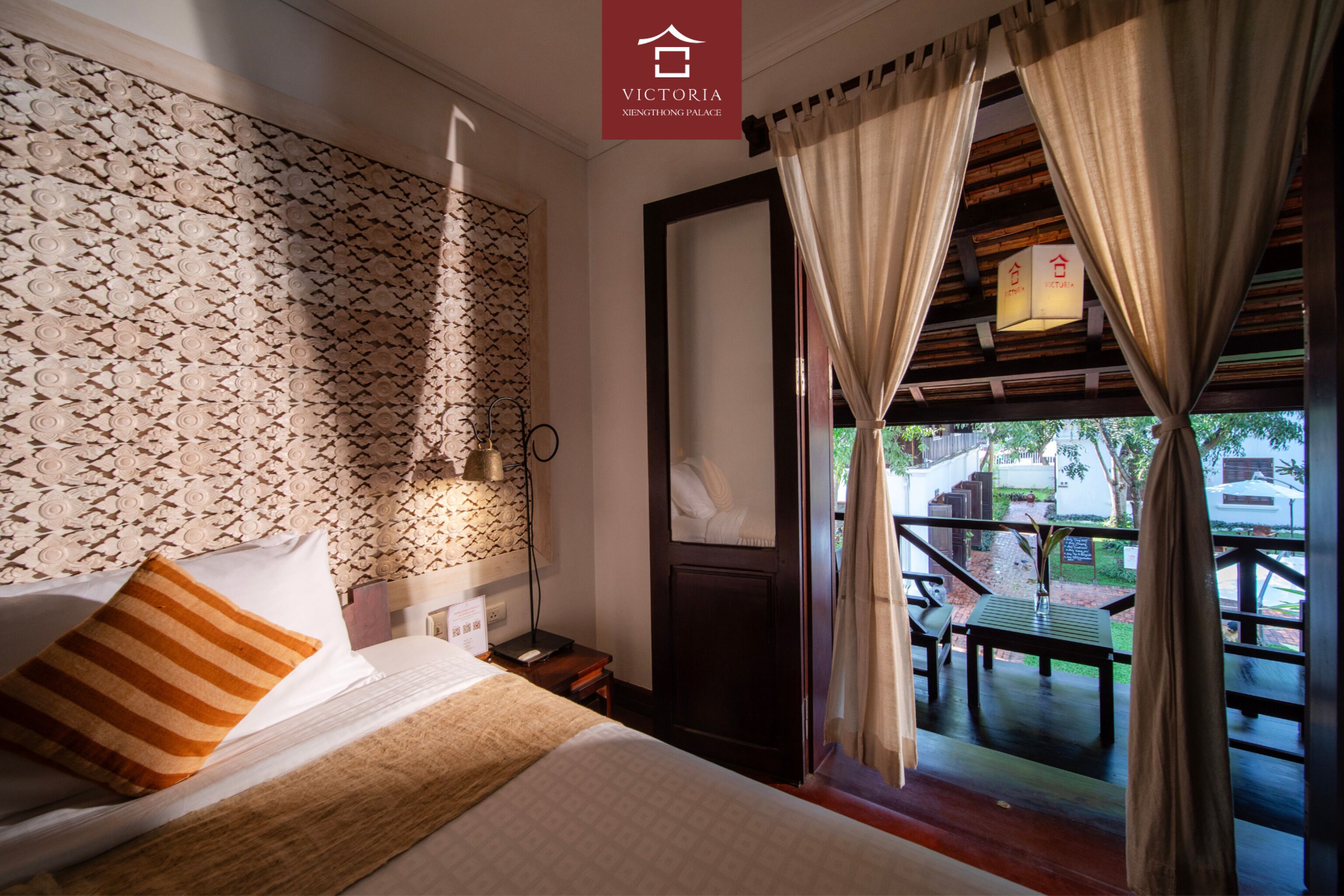 A hotel room with a patterned wallpaper, a bed with striped pillows, a nightstand, a desk, and a view of the outdoor patio area.