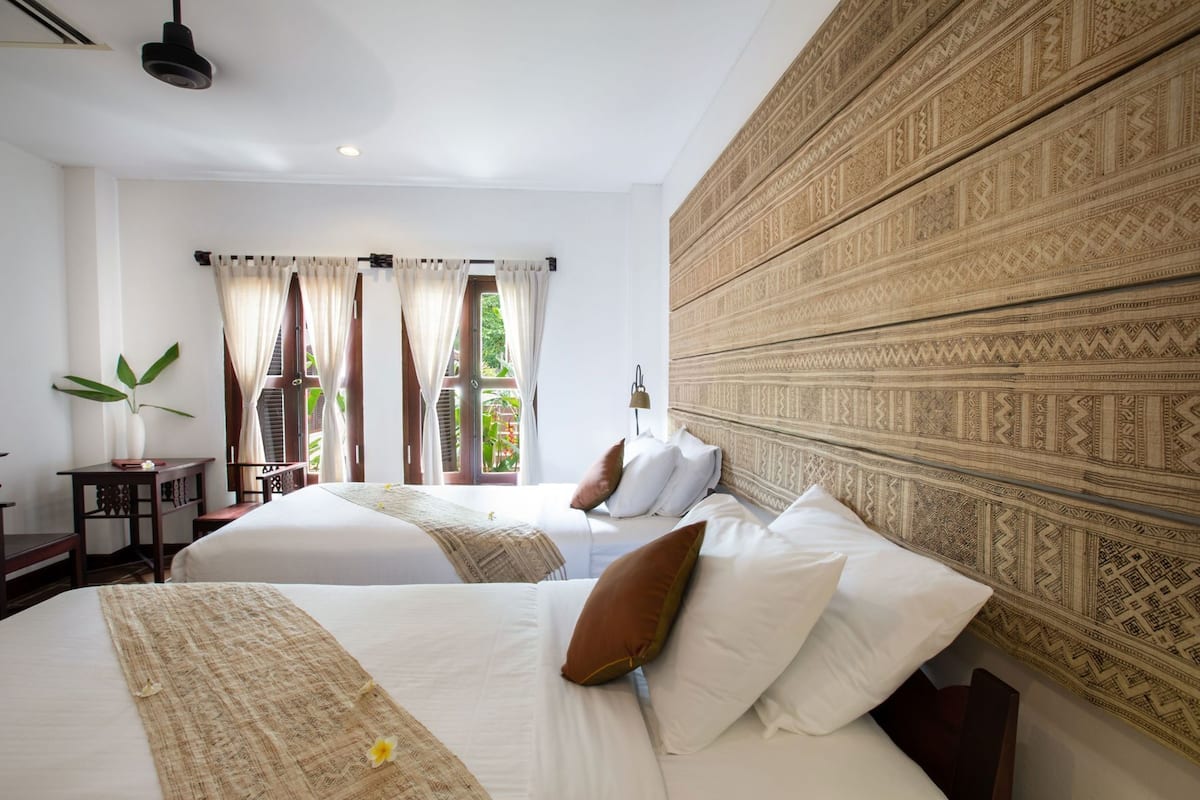 A hotel room with two beds, a patterned headboard, a window with curtains, a bedside table with a plant, and a lamp.