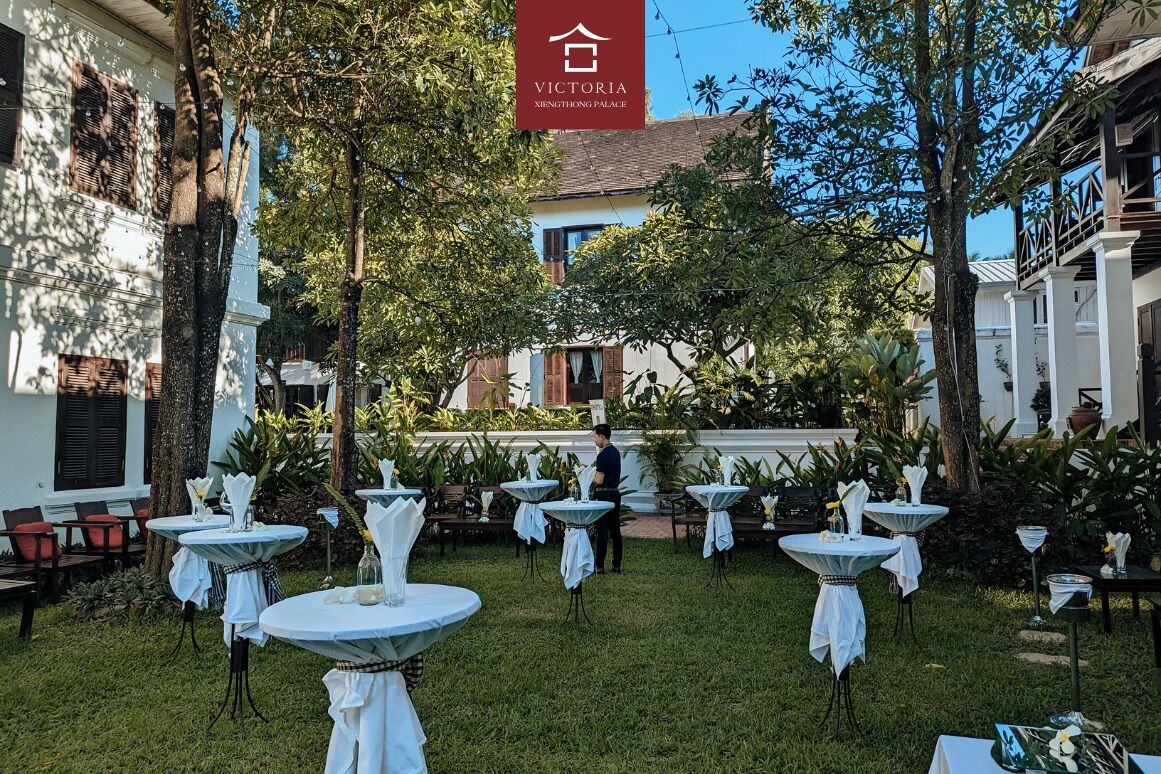 Un área de comedor al aire libre con mesas redondas cubiertas de manteles blancos, rodeada de follaje y árboles.