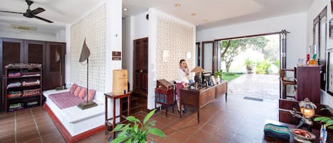 Un amplio vestíbulo de hotel con mostrador de recepción, una cama grande con colcha roja y un ventilador de techo.